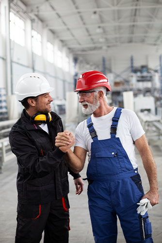 Zwei Fabrikarbeiter mit Schutzhelmen lächeln und schütteln sich die Hände in einer hell erleuchteten Industrieanlage. - © Shutterstock